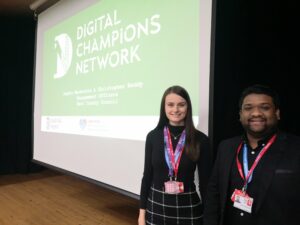 Two members of the Digital Kent Team stand in front of a projector screen with their logo.