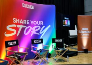 Image of BBC Share your Story Set. Behind is a rainbow-coloured poster with the logo on it. In front are three chairs facing forwards, and to the right is a pop up stand that says 'BBC bitesize careers'. 