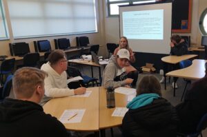 A presenter sits in front of a screen with task instructions. Four students sit around a table in front of her, listening and with worksheets in front of them. 